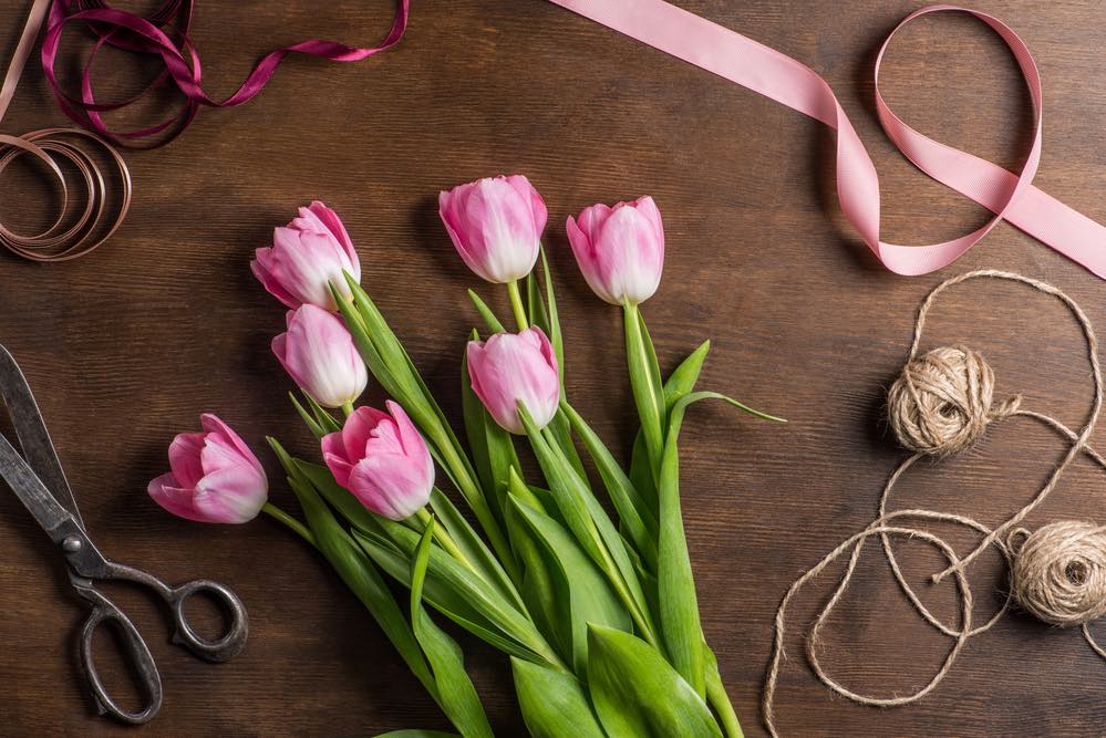 7 pink tulips, 2 balls of twine, 3 ribbons and a pair of scissors on a wooden table.