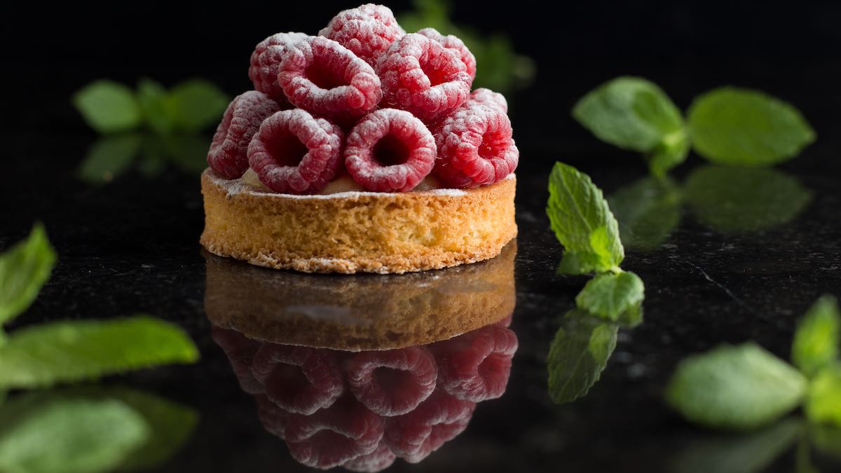 Raspberry tart dessert surrounded by tossed mint leaves on a dark, reflective flat surface