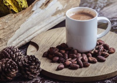 Sweet, hot cocoa drink in white cup among cocoa beans on wooden tabletop with pine cones